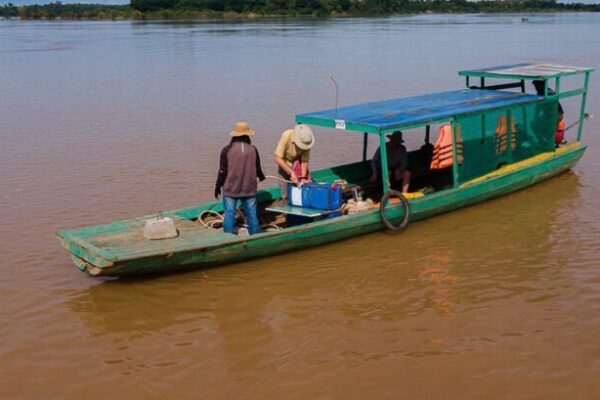 A boat with people standing and sitting on it while it floats in water. One person is bent over a rectangular container which is one part of a platform that will also have an acoustic receiver attached to it and be lowered into the water to float.