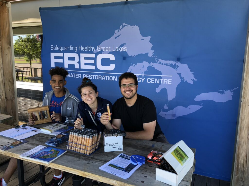 Three students sitting at a booth smile for the camera. They sit in front of a large sign with a picture of the Great Lakes.