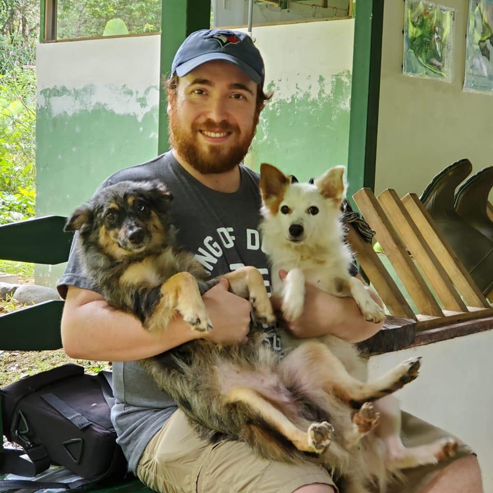Phillip Oelbaum A photograph in a Philip Oelbaum in a blue cap and t-shirt, holding two small dogs. One dog is black with brown patches, and the other is white with one brown ear.