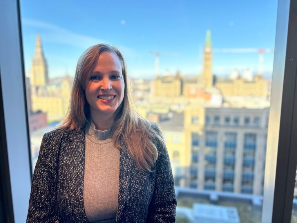 Sara Cannon A photo of Sara Cannon in a blouse and blazer, standing in front of a window with the Ottawa skyline in the background.